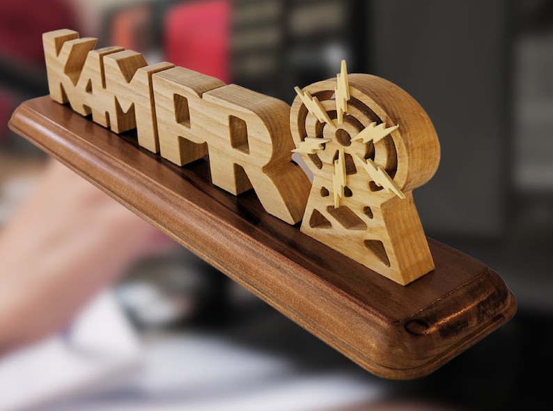 May include: Wooden desk sign with the name "KAMAR" in block letters, accompanied by a radio tower design. The sign is made of light-colored wood and mounted on a dark brown base. The radio tower has a circular design with lightning bolt accents.
