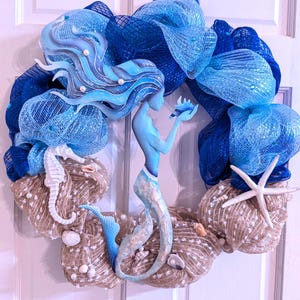 May include: A handcrafted mermaid-themed wreath featuring a central mermaid figure, seahorse, and starfish accents. The wreath incorporates shades of blue mesh and burlap, with seashells and pearl embellishments. This decorative wreath is designed for door or wall decor.