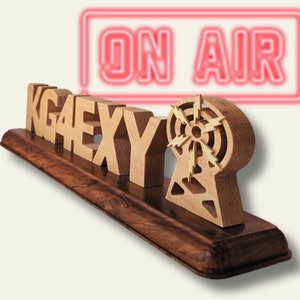 May include: A wooden radio station sign with the call letters KGAEXY, a radio tower, and the words "ON AIR" in a neon sign. The sign is made of light-colored wood and sits on a dark wooden base.