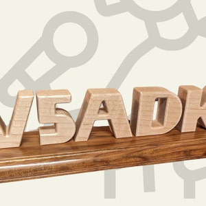 May include: Wooden desk sign featuring the letters "W5ADK" in a light wood color, mounted on a dark wood base. A wooden antenna is on the right. The sign is likely a personalized item or a decorative piece.