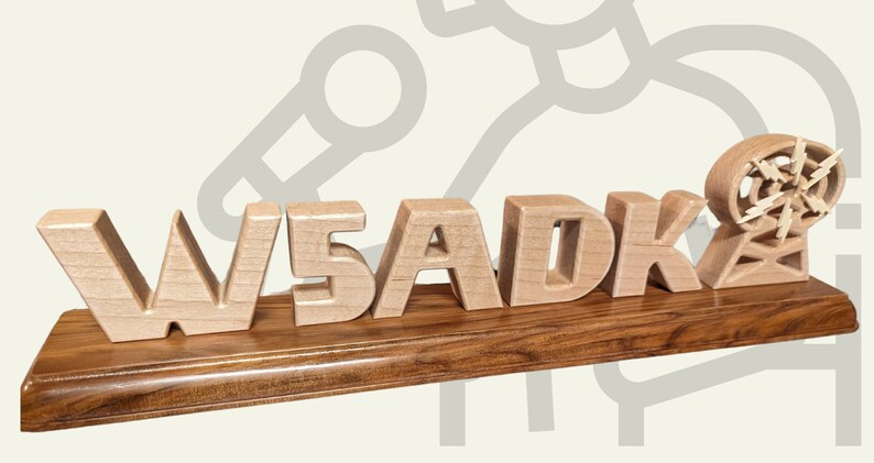 May include: Wooden desk sign featuring the letters "W5ADK" in a light wood color, mounted on a dark wood base. A wooden antenna is on the right. The sign is likely a personalized item or a decorative piece.