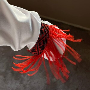 May include: Close-up of a white sleeve with a cuff adorned with black and red beaded patterns. Long red fringe hangs from the cuff, adding a decorative element to the garment. The sleeve is made of a smooth, white fabric.