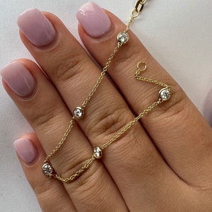 May include: A delicate gold chain bracelet with five small round clear stones. The bracelet is on a hand with light pink nail polish.