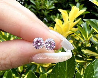 May include: A pair of silver stud earrings with round, light pink gemstones. The earrings are shown on a hand against a green and yellow leafy background.