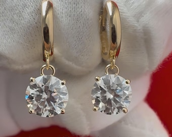 May include: A pair of gold hoop earrings with a dangling round clear stone. The earrings are on a white surface with the text "Natural Diamonds of NYC" below.