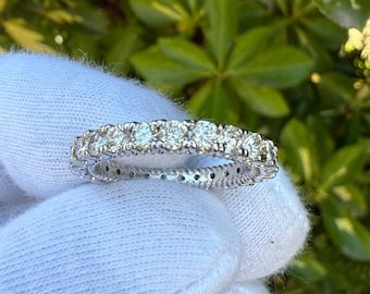 May include: A silver eternity ring with multiple round-cut diamonds. The ring is set in a delicate, detailed band. The diamonds sparkle brightly against the silver setting. The ring is shown against a background of green foliage.