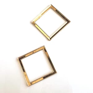 May include: Two gold-colored square frames are displayed against a white background. The frames are made of a thin, shiny metal and are positioned at a slight angle to each other. The frames are empty in the center.