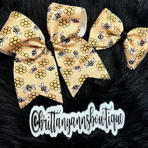 May include: Three hair bows with a yellow honeycomb and bee pattern. The bows are tied with black centers. The bows are on a black surface. The text "Brittanyannsboutique" is in a white and black cursive font.
