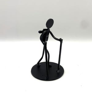 May include: A black metal sculpture of a person walking with a cane and a backpack. The figure is made of thin metal rods and is mounted on a circular base. The sculpture has a minimalist design.
