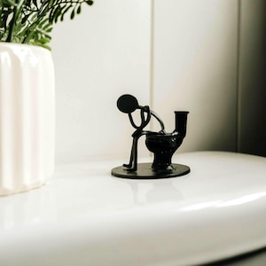 Könnte beinhalten: Schwarze Metallskulptur, die eine Person auf einer Toilette darstellt. Die Figur ist gebeugt, mit rundem Kopf und stäbchenartigen Gliedmaßen. Die Skulptur steht auf einem runden Sockel.