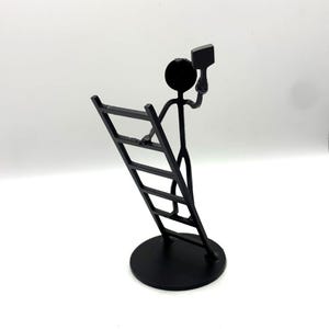 May include: A black metal sculpture of a stick figure climbing a ladder and holding a square object. The sculpture is on a round base and has a minimalist design. The background is white.