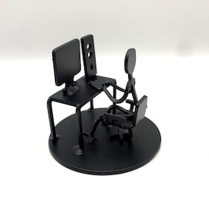 May include: A black metal sculpture depicts a person seated at a desk with a computer monitor and a second vertical element. The figure is seated in a chair, with arms extended toward the desk. The sculpture is mounted on a circular base.