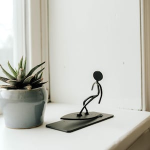 Op de afbeelding: Een zwarte metalen sculptuur van een stokfiguur die surft op een rechthoekig bord. De sculptuur staat op een wit oppervlak naast een vetplant in een grijze pot. De achtergrond is een witte muur en een raam.