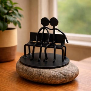 May include: A black metal sculpture of two figures sitting on a bench, mounted on a gray stone. The figures are stylized, with round heads and simple body lines. The bench has a minimalist design. The sculpture is placed on a wooden surface.