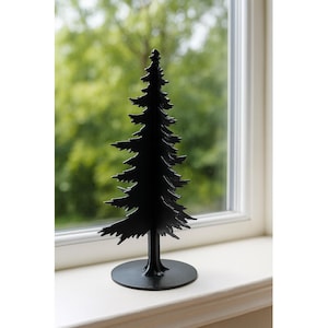 May include: A black metal decorative Christmas tree figurine. The tree has a detailed silhouette with layered branches and a circular base. It is placed on a white windowsill with a blurred green background.