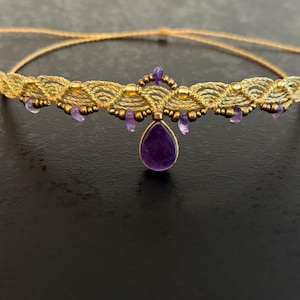 Op de afbeelding: Een goudkleurige macramé ketting met een centraal ontwerp met kleine paarse edelstenen en een grote druppelvormige paarse edelsteen hanger. De ketting wordt gepresenteerd op een donkere ondergrond.