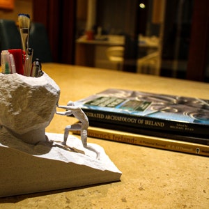 Op de afbeelding: Een witte 3D-geprinte bureau-organizer in de vorm van een rots met een klein figuurtje dat er tegenaan duwt. De organizer heeft een compartiment voor pennen en potloden. De organizer staat op een tafel met twee boeken, "Illustrated Archaeology of Ireland" en "Browne's Ireland".