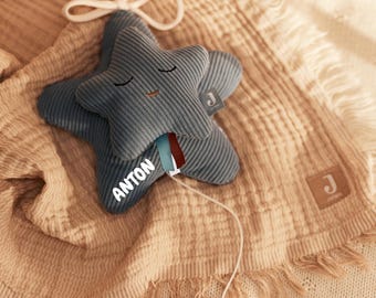 May include: A blue star-shaped baby toy with the name "ANTON" printed on it. The toy has a smiling face and a wooden ring attached to a string. It rests on a beige blanket with a fringed edge.