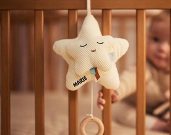 May include: Cream-colored star-shaped baby mobile with a smiling face and the name "Marie" printed on it. The mobile hangs from a wooden crib and features a wooden ring at the bottom. The star has a textured, ribbed design.