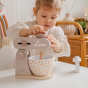 Könnte beinhalten: Ein hellbrauner Spielzeug-Standmixer aus Holz mit einer weißen Schüssel und einem braunen Holzgriff. Der Mixer hat ein weißes Etikett mit der Aufschrift "Lara". Die Schüssel hat ein braunes und weißes Punkte-Muster.