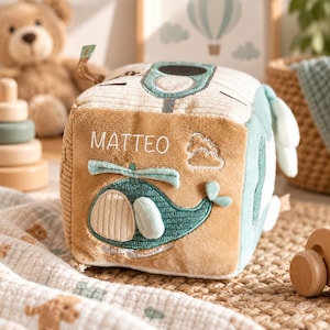 Könnte beinhalten: Ein weicher, quadratischer Baby-Aktivitätswürfel aus Plüsch in den Farben Beige und Salbeigrün. Die Vorderseite zeigt einen gestickten türkisfarbenen Hubschrauber und den Namen MATTEO in weißer Schrift sowie ein kleines gesticktes Wolkensymbol.