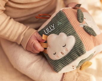 May include: A soft, multi-coloured baby activity cube with the name "LILLY" printed on it. The cube features a cloud, star, and leaf design. The cube is made of corduroy and other soft fabrics.