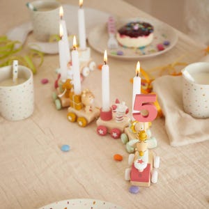 Puede incluir: Una escena festiva de cumpleaños con un tren de madera que lleva velas y una vela con el número 5. Los vagones del tren presentan diseños de animales en colores pastel. Velas blancas encendidas y una mesa con golosinas y confeti.