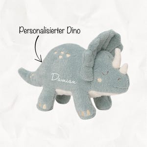 Könnte beinhalten: Ein weiches, hellblaues Triceratops-Dinosaurier-Spielzeug mit weißen Hörnern und Akzenten. Der Name "Damian" ist auf der Seite gestickt. Der Text "Personalisiert Dino" befindet sich darüber.