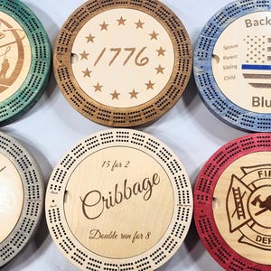 May include: Six wooden cribbage boards with different designs. The boards are circular and have a wooden center with a design and a wooden outer ring with holes for scoring. The designs include a fish, the year 1776, a thin blue line flag, a bulldog, a cribbage board, and a fire department logo.
