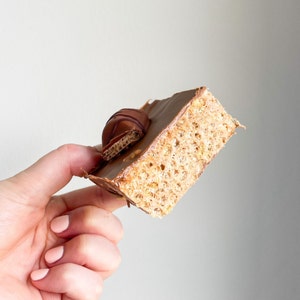 May include: A chocolate-covered rice crispy treat with a chocolate candy on top. The treat is being held by a hand.