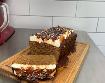 May include: A loaf of cake with white frosting and chocolate drizzle, topped with nuts and blue edible flowers, on a wooden cutting board.