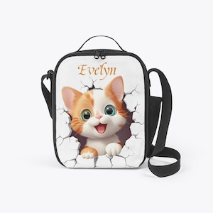Könnte beinhalten: Weiße Lunchtasche mit schwarzem Griff und Schultergurt. Die Tasche zeigt ein Cartoon-Katzen-Design mit orange-weißem Fell, grünen und blauen Augen und dem Namen "Evelyn" in goldener Schrift.