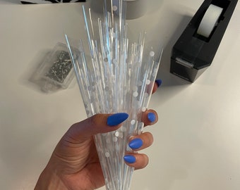 May include: A hand holding a bundle of clear, cone-shaped cellophane wrappers with white polka dots. The wrappers have silver wire at the bottom. A lit candle, tape dispenser, and a zebra-print bag are in the background.