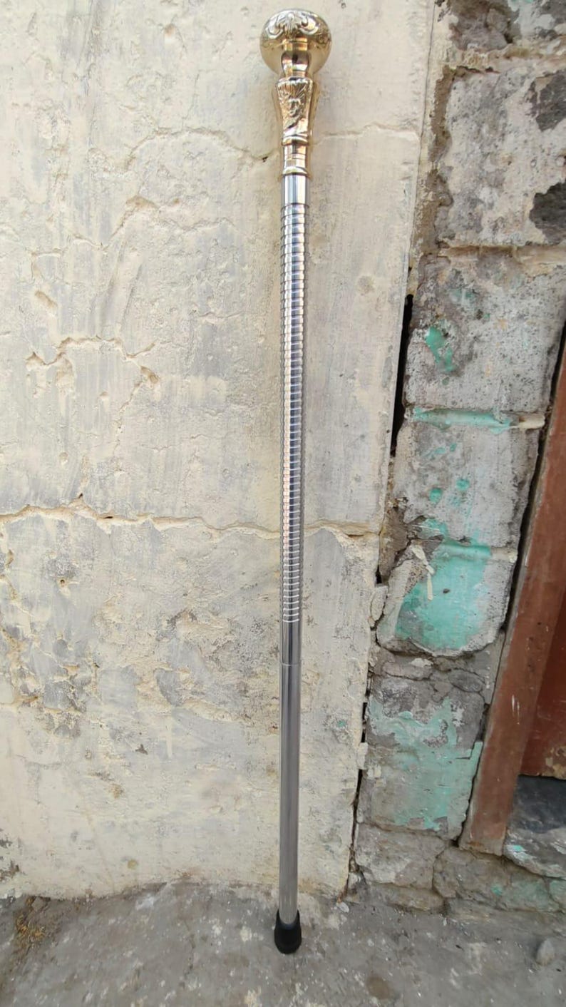 Puede incluir: Un bast&oacute;n de plata con una parte superior dorada con un dise&ntilde;o ornamentado. El bast&oacute;n tiene una punta de goma negra.