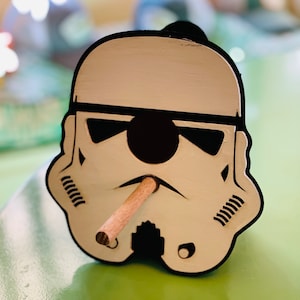 May include: A wooden Stormtrooper head-shaped ashtray with a cigar. The ashtray is painted white with black accents, including sunglasses and a black outline. The cigar is light brown and placed in the mouth of the Stormtrooper.