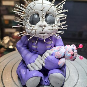 May include: A handmade, seated figurine with a white head adorned with silver pins. The figure wears a purple outfit and holds a small, colorful plush toy. The figurine has gray eyes and a neutral expression.
