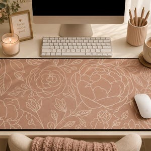 May include: A rectangular desk mat featuring a dusty rose background with a white line art pattern of large blooming flowers and leafy stems. The mat has a black border and is positioned on a desk surface with a white computer mouse resting on it.