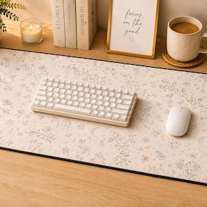 May include: A rectangular desk mat featuring a light beige background with a repeating pattern of small pink flowers, green foliage, and crescent moons. The mat has a black stitched edge and supports a white mechanical keyboard and a white computer mouse.