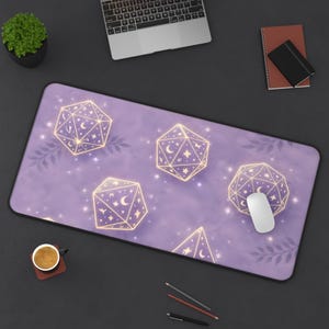 May include: A rectangular lavender mouse mat with a repeating pattern of gold geometric dice with celestial designs. A white computer mouse rests on the mat. The background includes a laptop, a small plant, and a cup of coffee.