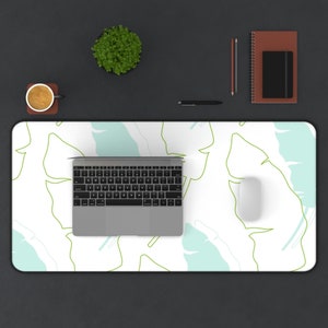Könnte beinhalten: Eine weiße Schreibtischunterlage mit einem abstrakten Blatt-Muster in Grün und Blau. Auf der Schreibtischunterlage befinden sich ein Laptop, eine kabellose Maus und eine Tasse Kaffee.