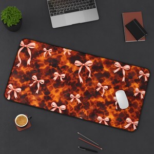 Puede incluir: Alfombrilla de escritorio rectangular con un diseño de carey y lazos rosas. La alfombrilla tiene un borde negro y se muestra con un ratón de ordenador, un portátil y otros suministros de oficina.