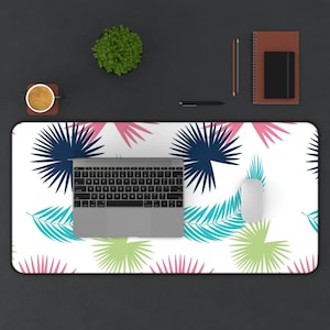 Könnte beinhalten: Eine weiße Schreibtischmatte mit einem tropischen Blatt-Muster in den Farben Rosa, Blau und Grün. Ein Laptop, eine kabellose Maus und eine Tasse Kaffee befinden sich auf der Matte.