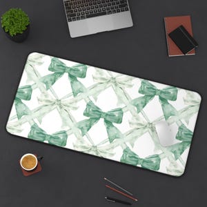 May include: A rectangular mouse mat featuring a repeating pattern of green and white watercolour bows. A computer mouse sits on the mat. The mat is on a dark surface with a laptop, notebook, and a cup of coffee.