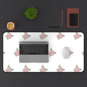 May include: A white desk mat featuring a floral pattern of pink roses and green leaves. A laptop, white mouse, and other office supplies are on the black desk. A small plant and a cup of coffee are also visible.