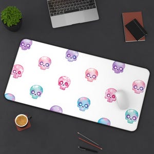 May include: White rectangular mouse mat with a repeating pattern of watercolour skulls in pink, blue, and purple. A computer mouse rests on the mat. The mat is on a dark surface.