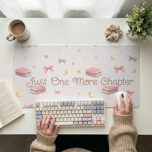 May include: A white desk mat with a repeating pattern of books, bows, stars, and moons. The text "Just One More Chapter" is in the center. A keyboard and mouse are on the mat, along with a cup of coffee and a plant.