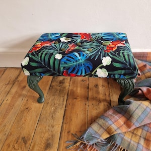 May include: A rectangular footstool with a tropical floral print in blue, red, and white on a black fabric. The dark green legs are curved. The stool is on a wooden floor. The dimensions are approximately 60 cm in length.
