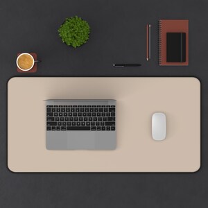 May include: A beige desk mat with a laptop, a wireless mouse, and a cup of coffee on it. There is a potted plant, a notebook, and two pens on the black surface surrounding the mat.