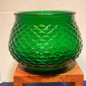 Könnte beinhalten: Eine leuchtend grüne Glasschale mit einem strukturierten, schuppenartigen Muster. Die Schale hat eine abgerundete Form und steht auf einem kleinen, quadratischen Holzständer. Das Glas hat eine glänzende Oberfläche und reflektiert das Licht.