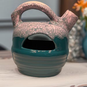 May include: A ceramic pitcher with a handle and spout. The top is pink with a textured pattern, transitioning to teal. The base is teal with horizontal ridges. The pitcher has a large opening in the front.